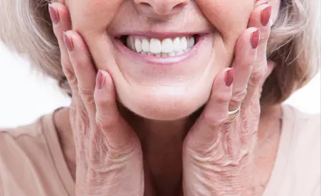 Eine ältere Frau mit strahlendem Lächeln hält ihre Wange, symbolisch für Freude und Zufriedenheit mit ihrer Zahngesundheit.